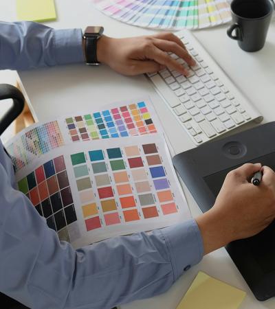 设计师在白色办公桌上使用绘图板和手写笔同时参考色样表的高角度视图。工作区包括无线键盘、桌面显示器、咖啡杯和一套彩色粉彩。