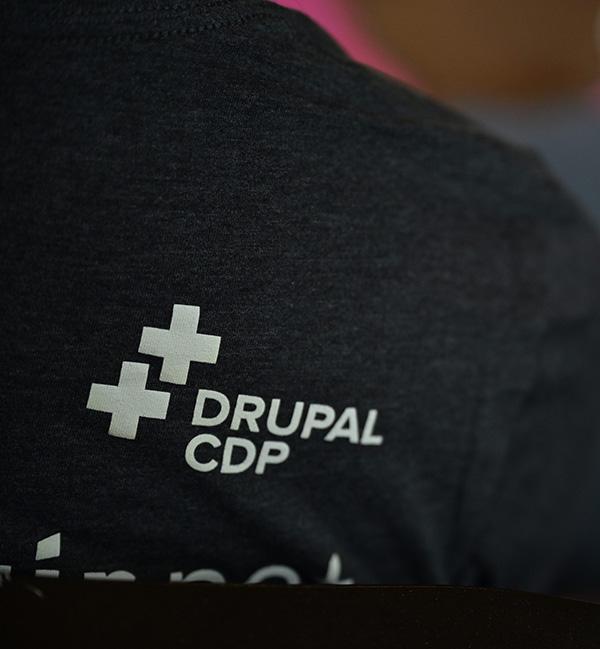 深灰色杂色 T 恤背面特写，带有白色“DRUPAL CDP”徽标。该徽标在文本旁边包含两个重叠的加号。背景柔和模糊。